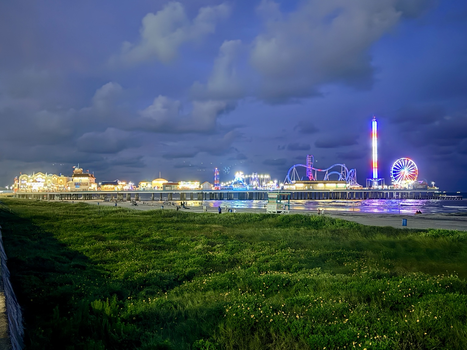 Galveston beach and illuminated Pleasure Pier rides beneath a cloudy evening sky.