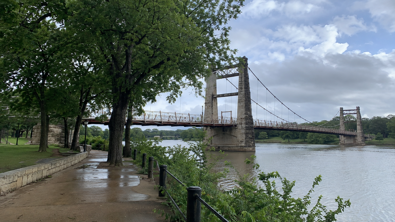 Waco Suspension Bridge spanning the Brazos River beside a tree-lined riverside path.
